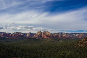 Photo is overlooking a valley in Arizona with red rock, pine trees and a blue, partly cloudy sky